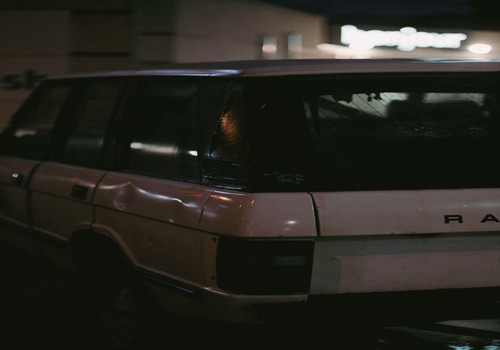 Old white SUV with visible dents on the rear side, parked under dim lighting at night.









