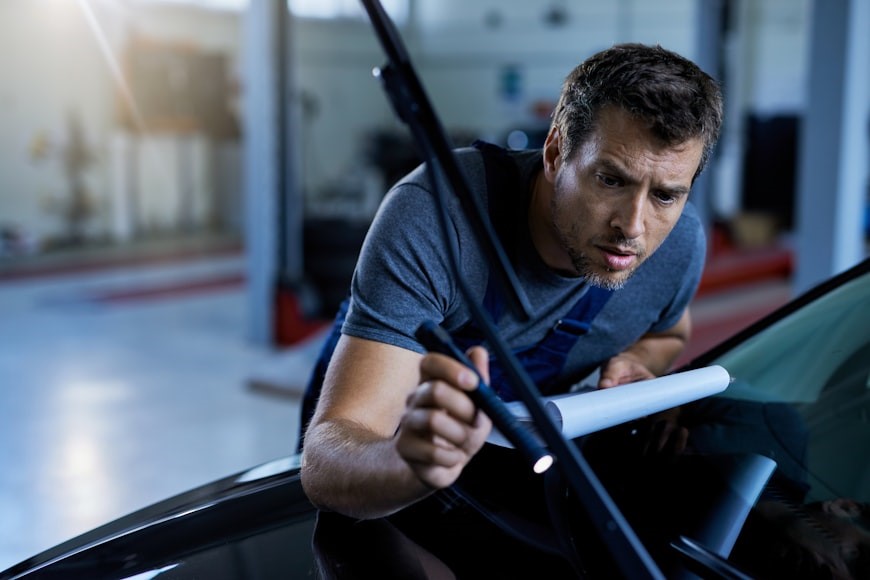 Mechanic carefully inspecting car windshield damage under bright workshop light.