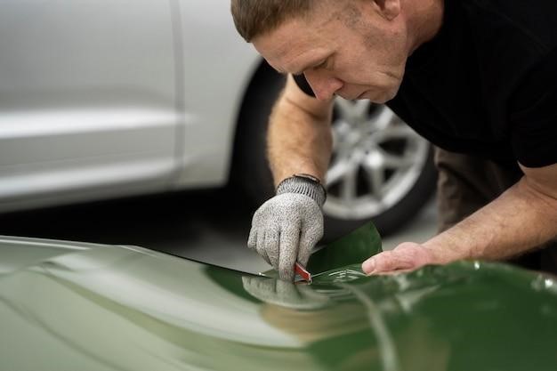 Man carefully applying green vinyl wrap to a car’s surface, inspecting for hidden damage.