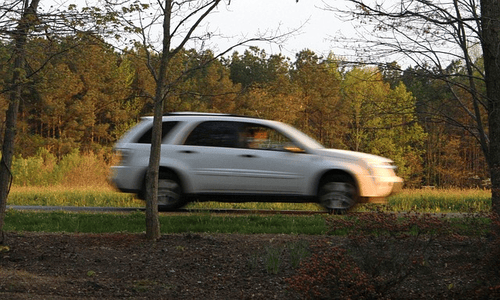 White SUV driving fast on a rural road with blurred motion and trees in the background.