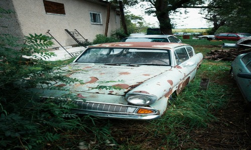 Old car with rust and peeling paint parked by house in overgrown grass.